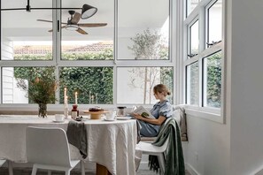 A dining area with a table set for two, a ceiling fan, and large windows with a view of greenery.