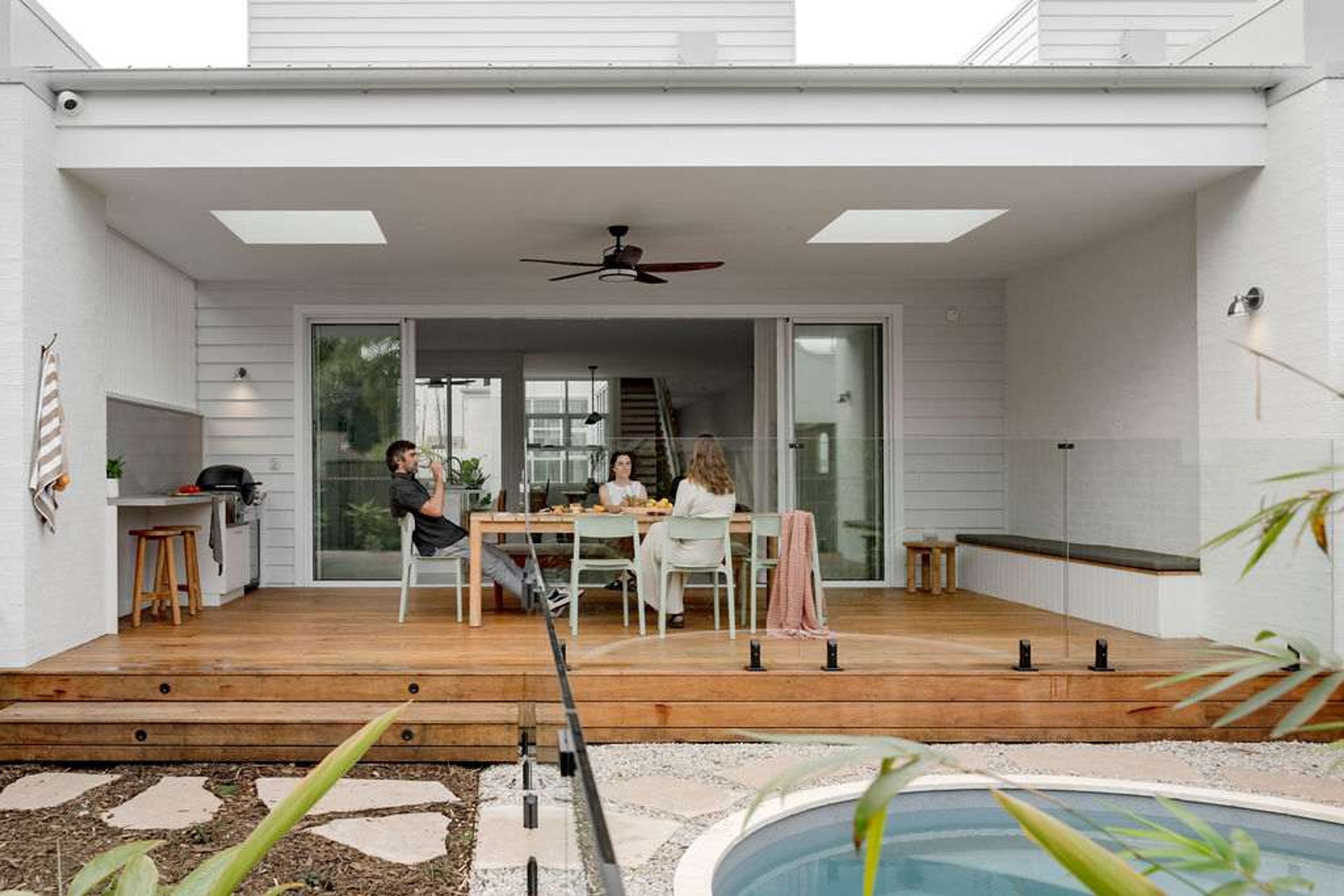 Una casa moderna con una terraza de madera, piscina y un comedor con tres personas.
