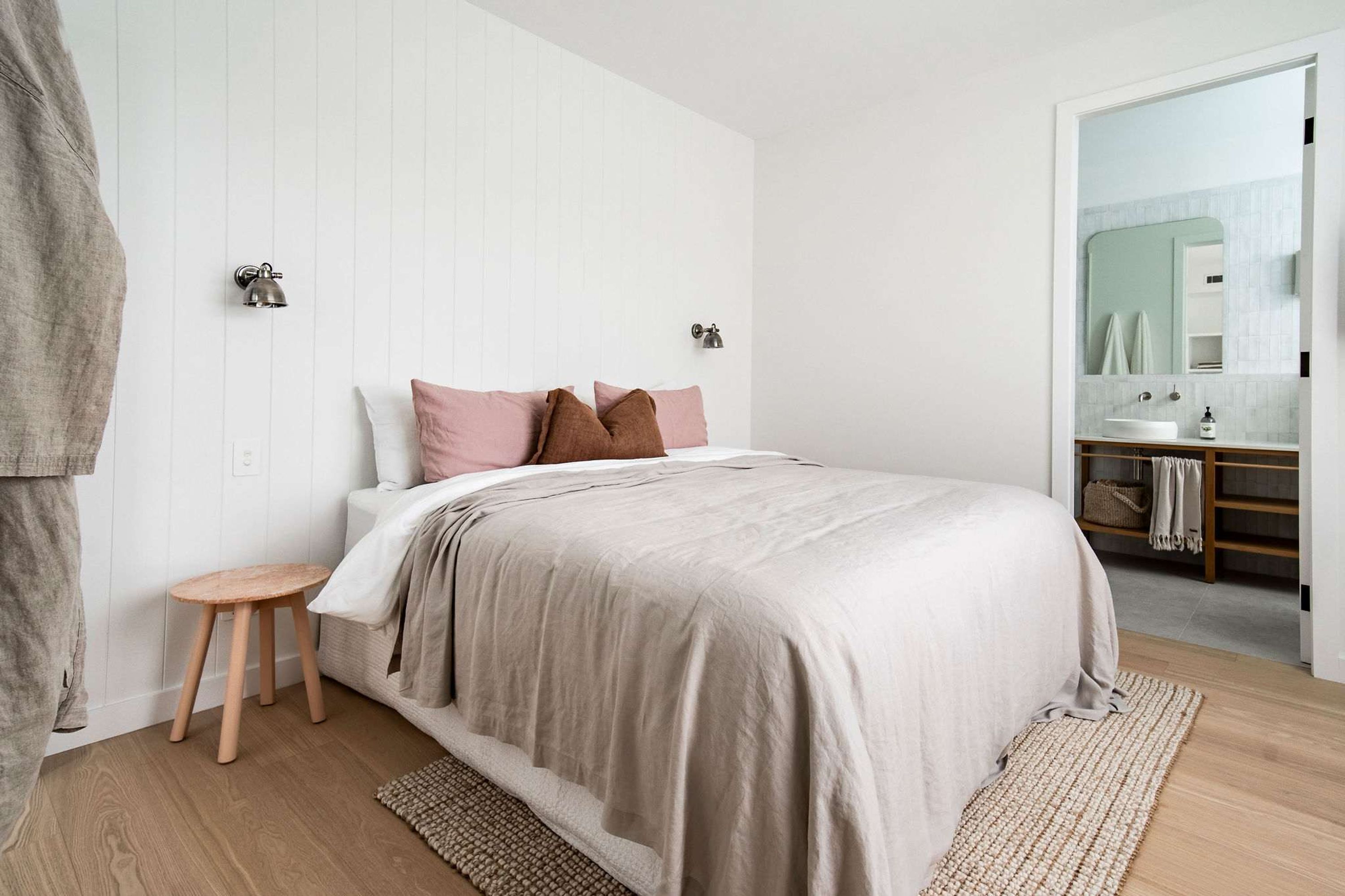 A bedroom with a bed, a small wooden stool, a mirror, and a bathroom visible through an open door.