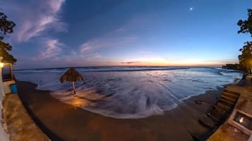 Una playa con un paraguas de paja, escalones de madera y vista al océano.