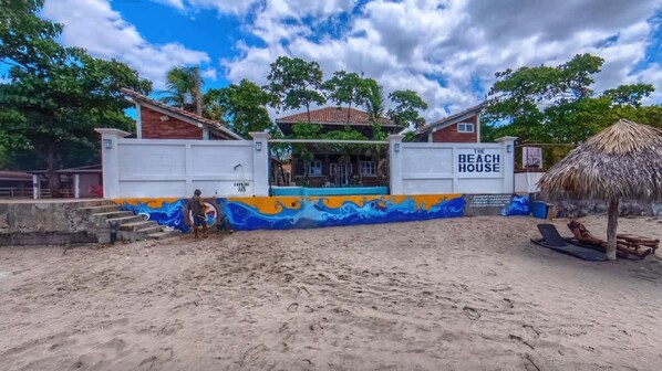 Une maison de plage ornée d’une fresque colorée, avec une enseigne indiquant « The Beach House », et un parasol en chaume planté dans le sable.