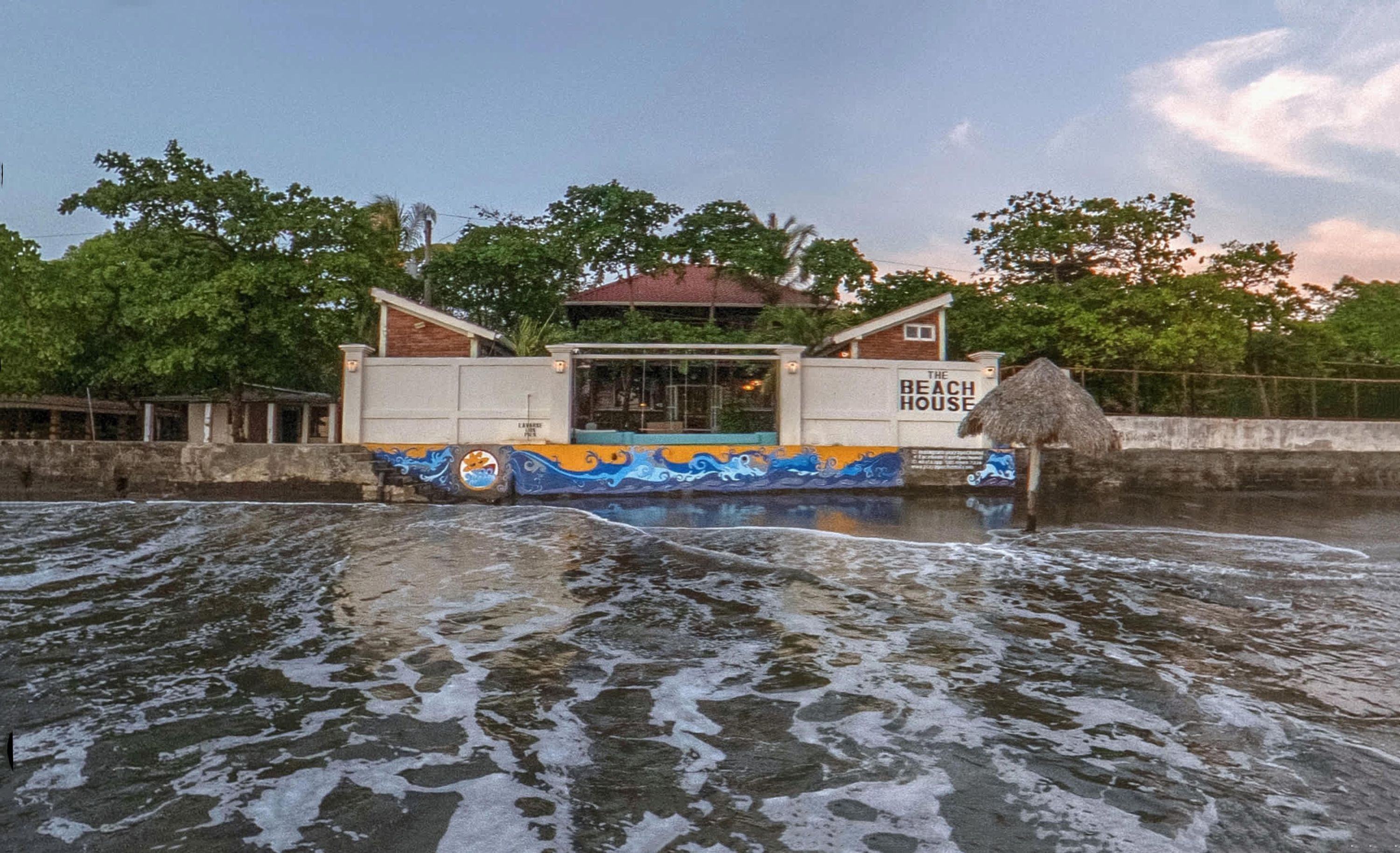 Un edificio con techo de paja, identificado como "The Beach House", situado junto a una masa de agua con olas.
