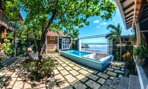 A pool area with a view of the ocean, surrounded by a garden and a building with a red roof.