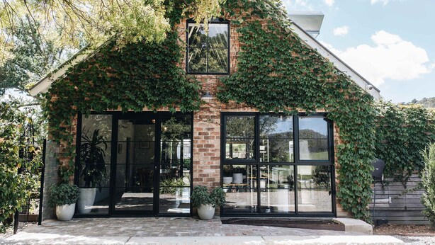 A house with a brick facade partially covered in ivy, large glass windows, and a visible patio area with potted plants. - Moraea Farm Berry - by Linnaeus Collection (Berry)
