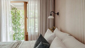 A bedroom with a bed, white and gray pillows, a wall-mounted lamp, and a view of greenery through a window. - Moraea Farm Berry - by Linnaeus Collection (Berry)