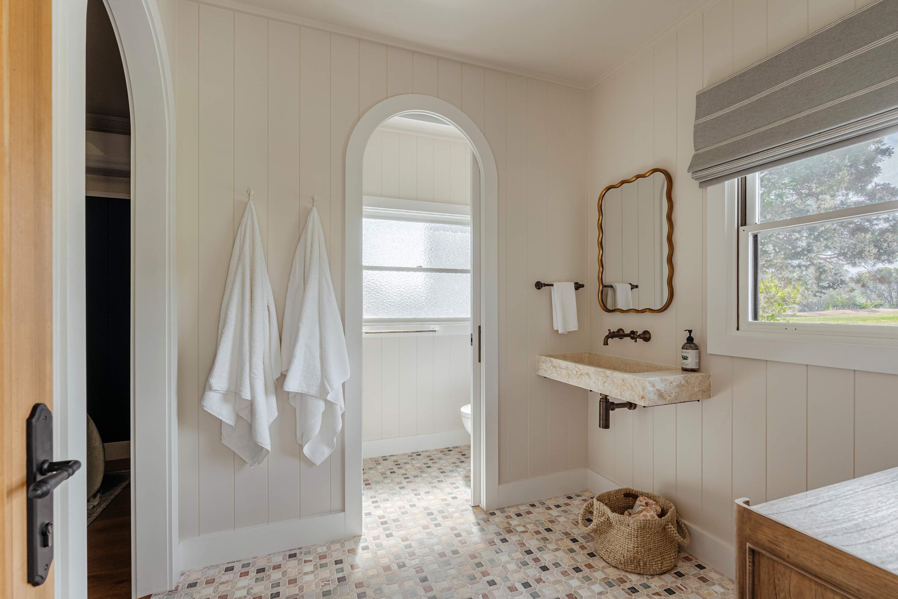 A bathroom with a white tiled floor, a wooden door, a white sink with a mirror, and a window with a view of trees.