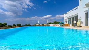 A clear blue swimming pool with lounge chairs and umbrellas, surrounded by a white building with large windows and a view of the sky. - VIP Villa Panoramic Seaview Close to Beaches (Ermionida)