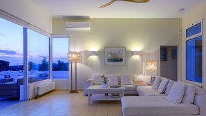 A modern living room with a large sofa, a coffee table, and a floor lamp. There is a ceiling fan, a window with a view, and a picture on the wall. - VIP Villa Panoramic Seaview Close to Beaches (Ermionida)