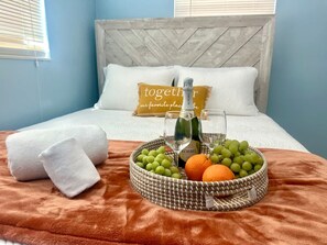 Un lit bien fait, avec une tête de lit, une table de chevet sur laquelle se trouvent une bouteille et des verres, et un panier de fruits posé sur le lit.