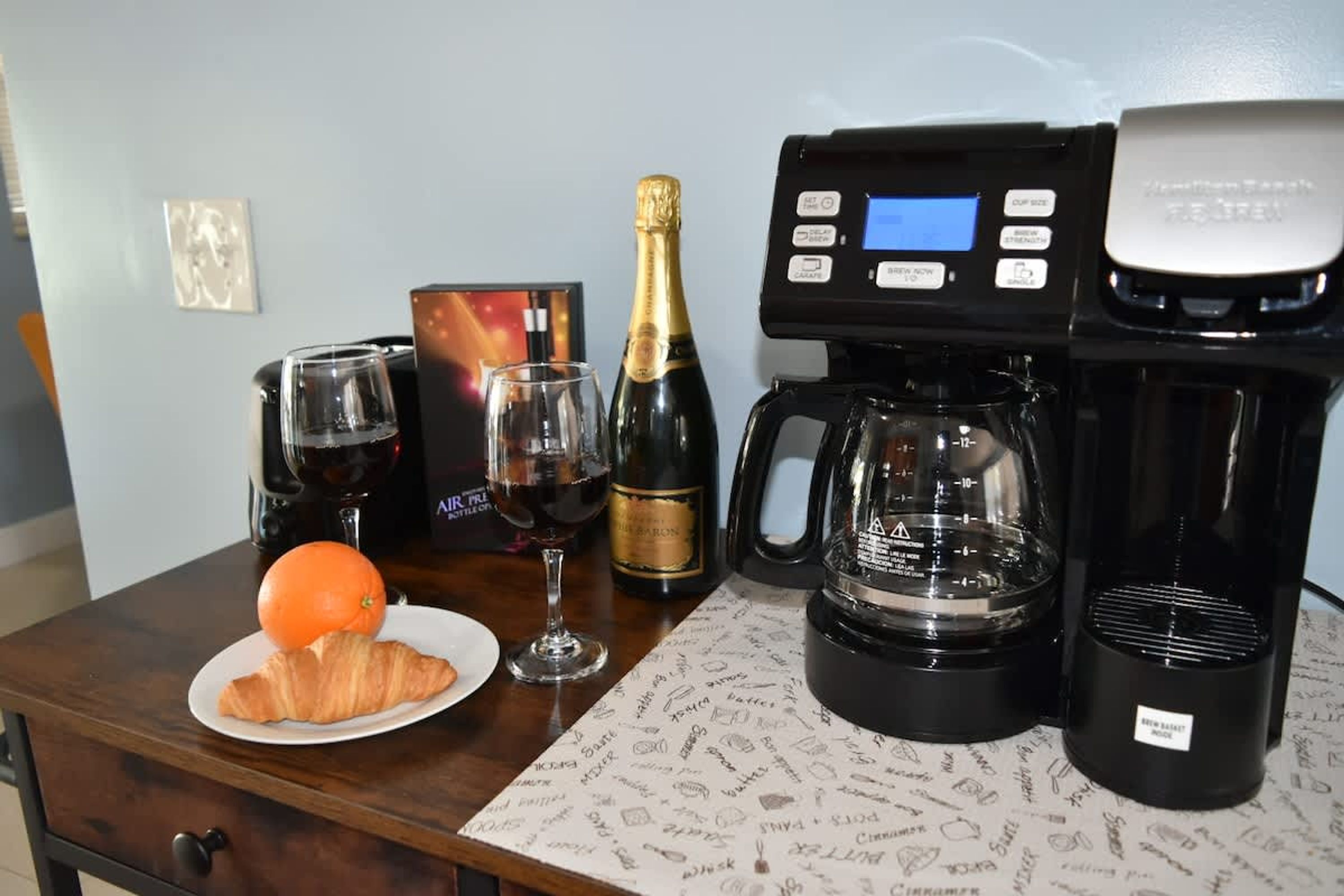 A table with a coffee maker, a glass of red wine, a bottle of champagne, a glass, and a croissant on a placemat.