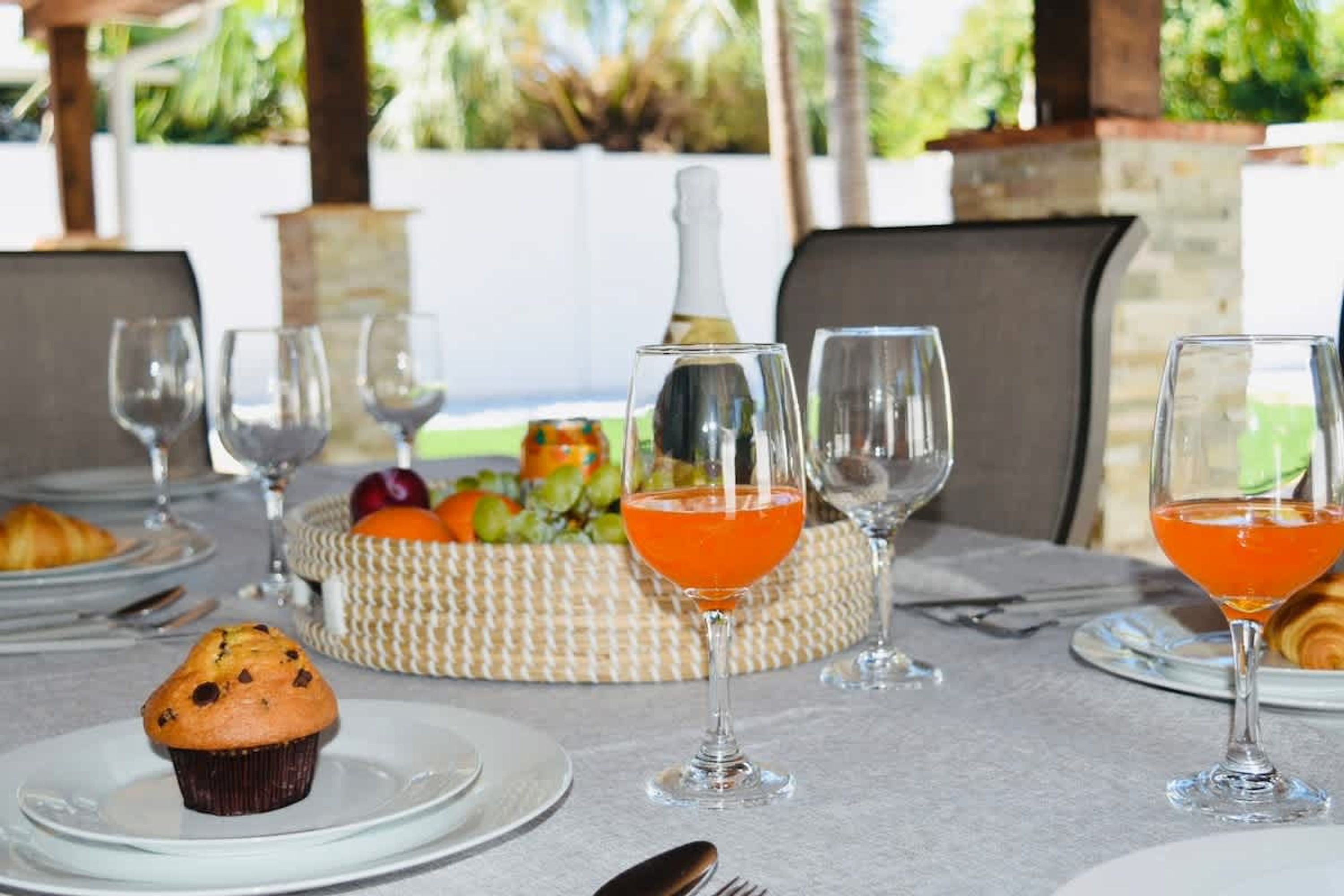 A table set for a meal with plates, glasses, a muffin, a fruit basket, and a bottle.