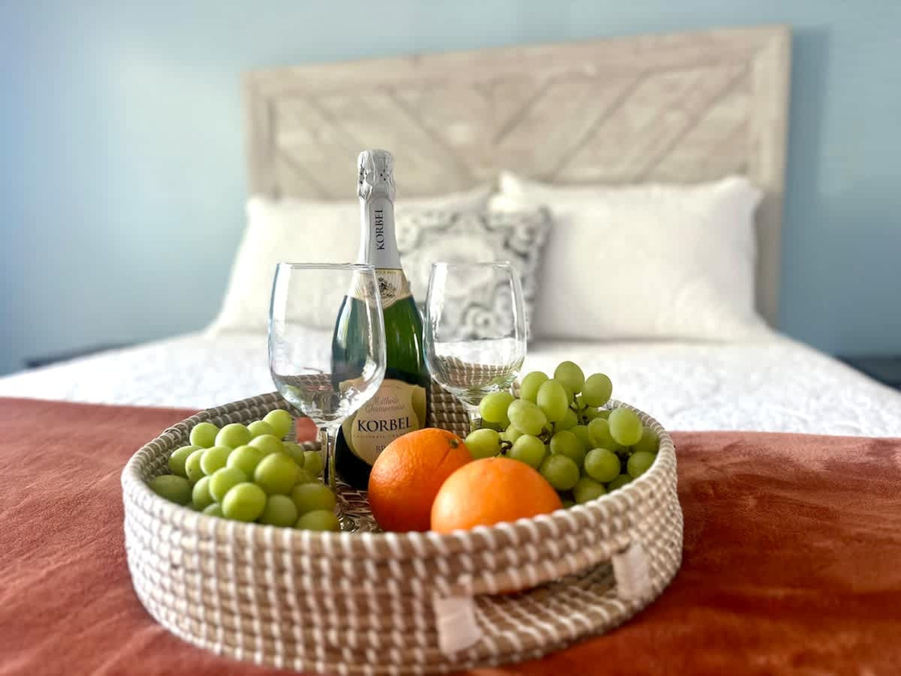 Een bed met een wit hoofdeinde, een rieten mand met groene druiven, een sinaasappel en een fles Korbel wijn.