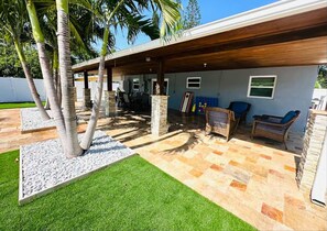 An outdoor patio area with a covered seating area, stone flooring, and palm trees.
