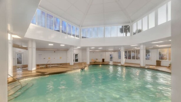 An indoor swimming pool with clear water, surrounded by white walls and columns, and large windows allowing natural light. - Good Vibes - The Affordable Beach Vacay (Gulf Shores)