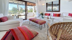 Ein modernes Wohnzimmer mit einer großen Schiebeglasdoor zum Balkon, weißen Sofas mit bunten Kissen, einem weißen Couchtisch und einem Korbstuhl.
