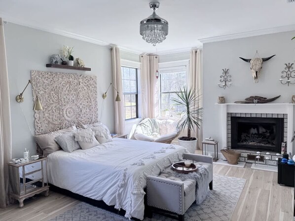 A bedroom with a large bed, a bedside table, a chair, a fireplace, and a chandelier.