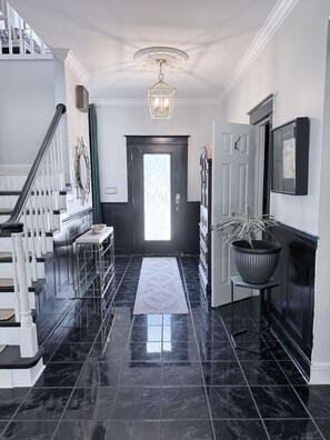 A hallway with a black tiled floor, a white bench, a staircase with white railings, a black door with a glass panel, and a potted plant.