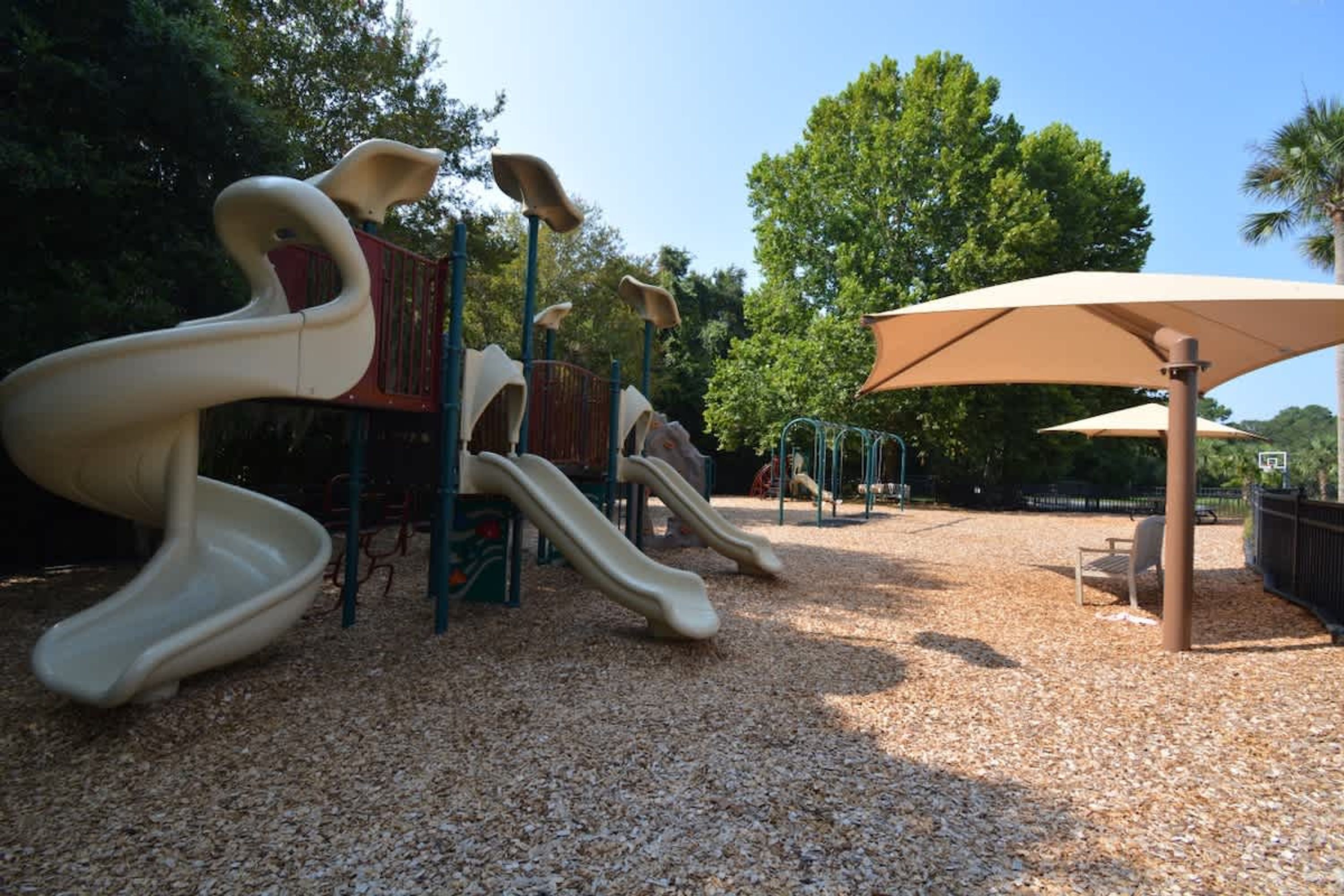 Un'area giochi con scivoli e strutture per arrampicarsi, circondata da alberi e riparata da un grande ombrellone.