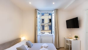 Habitación de hotel con cama, escritorio, silla, televisor y ventana con cortinas.