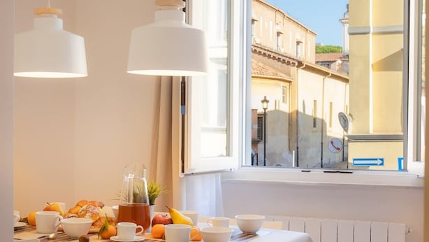 Una mesa de desayuno con cruasanes, frutas y tazas de café junto a una ventana con vistas a edificios.