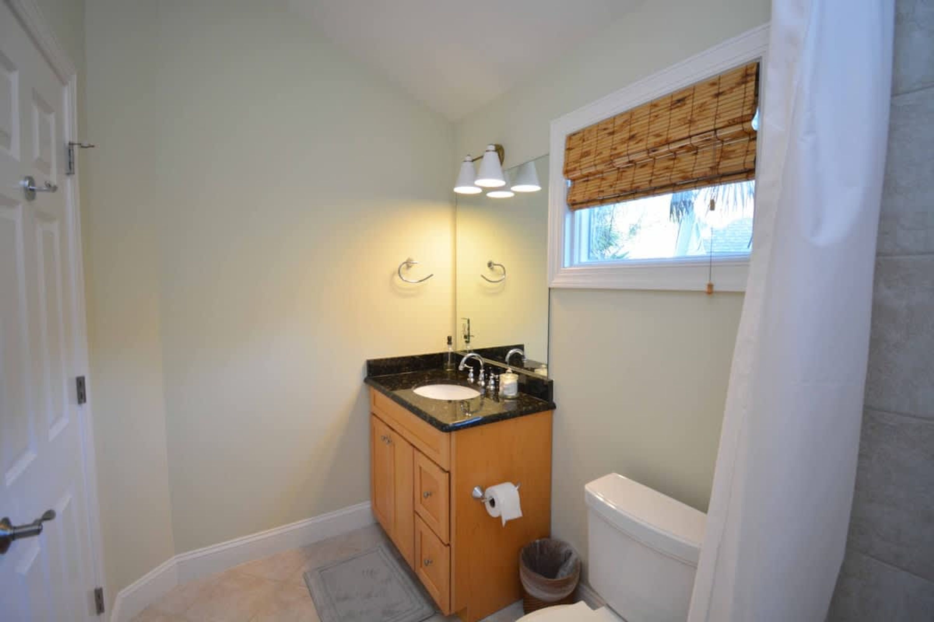 A bathroom with a wooden vanity, a white toilet, a window with a bamboo blind, and a small trash bin.