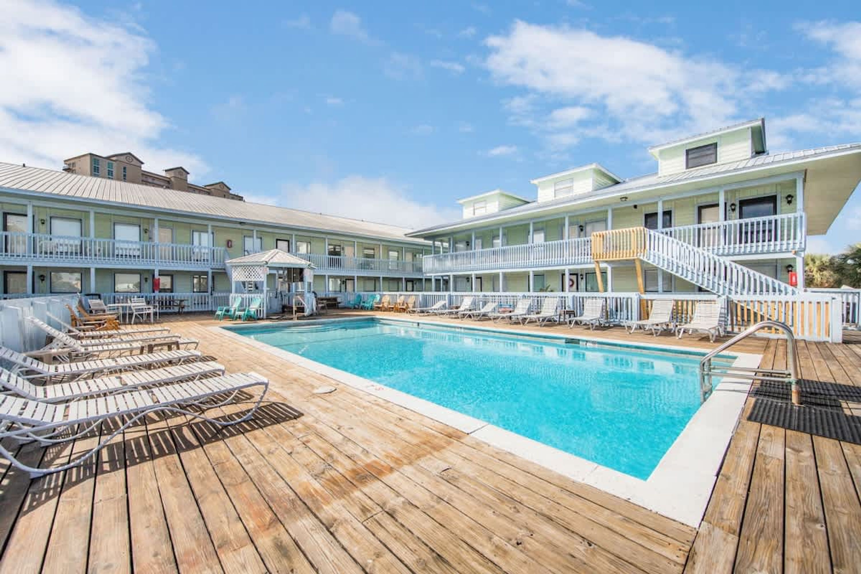 A pool area with wooden deck, lounge chairs, and a building with balconies.