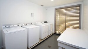 Une buanderie équipée de trois machines à laver et de trois sèche-linge blancs, d’une petite table et d’une fenêtre avec des stores.
