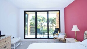 Une chambre dotée d’une grande porte-fenêtre coulissante donnant sur un balcon extérieur, avec un lit, une commode, une chaise et une lampe.