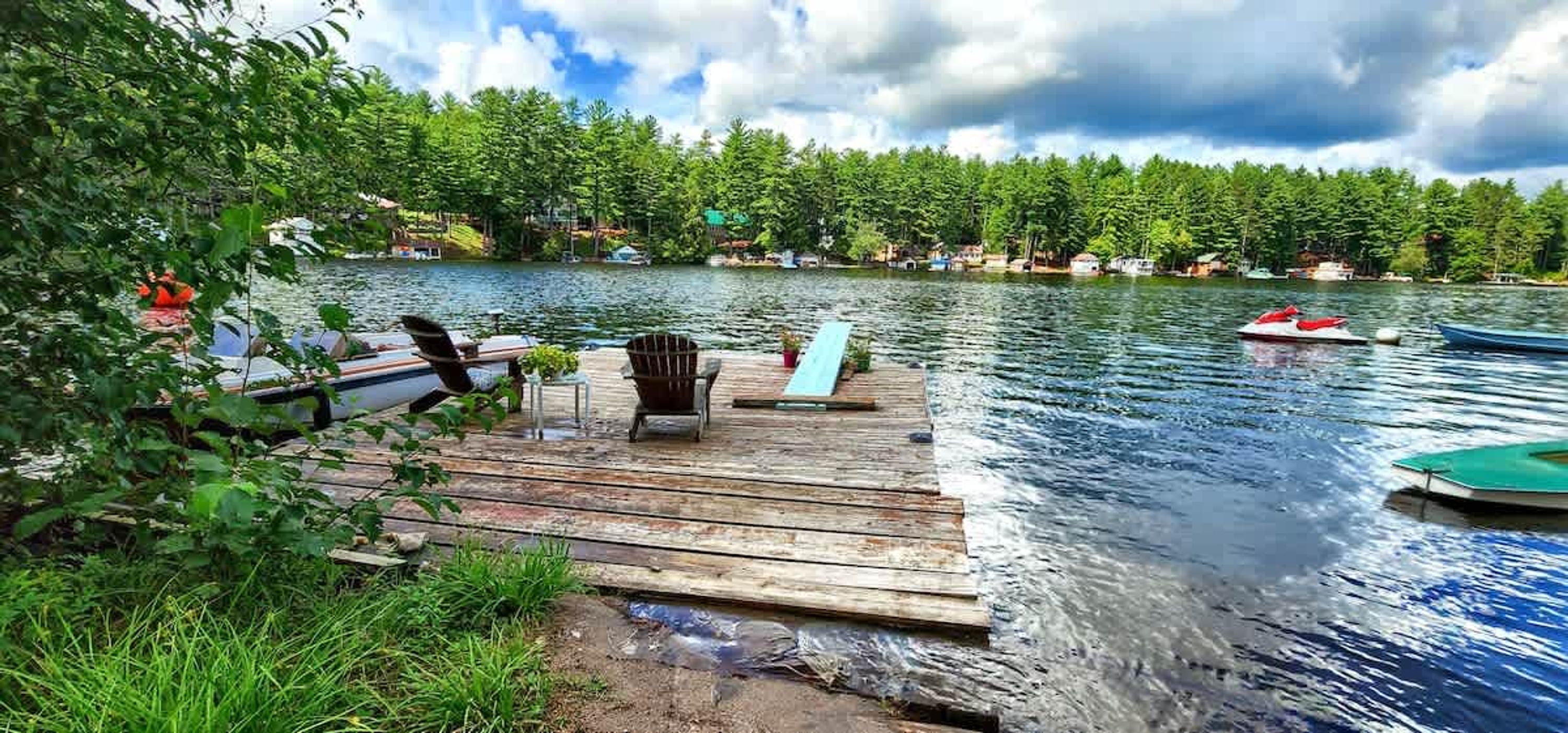 A wooden dock extending into a lake with boats, surrounded by trees and houses in the background.