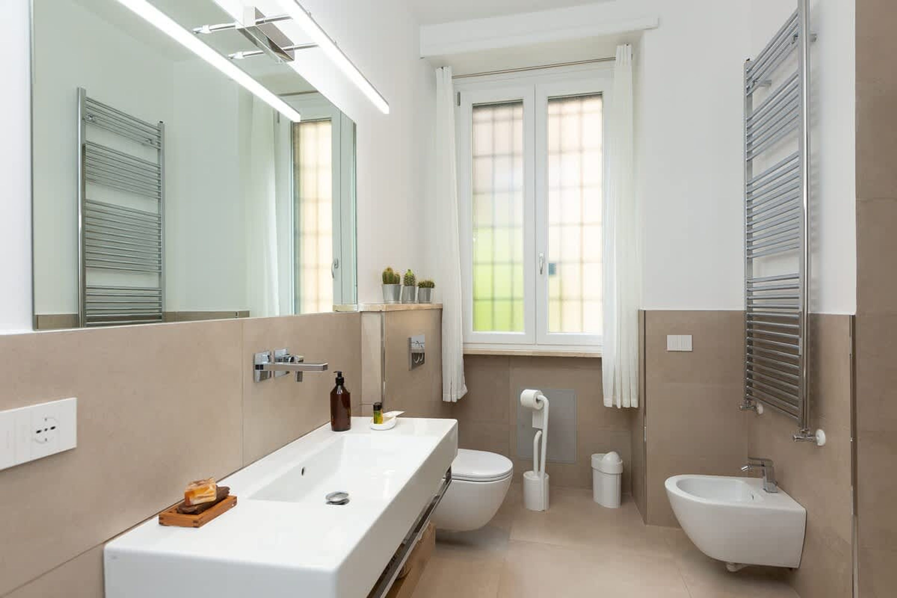 A modern bathroom with a large sink, toilet, bidet, and a window with curtains.