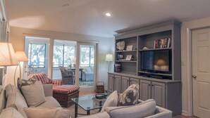 A living room with a sofa, armchairs, a glass coffee table, a television, and a bookshelf. - Best Value in Seabrook - Heron Run (Seabrook Island)
