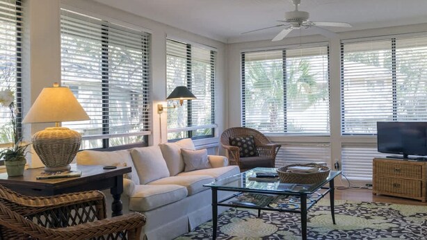 A spacious living room with a sofa, a glass coffee table, wicker chairs, a television, and a ceiling fan. - Best Value in Seabrook - Heron Run (Seabrook Island)