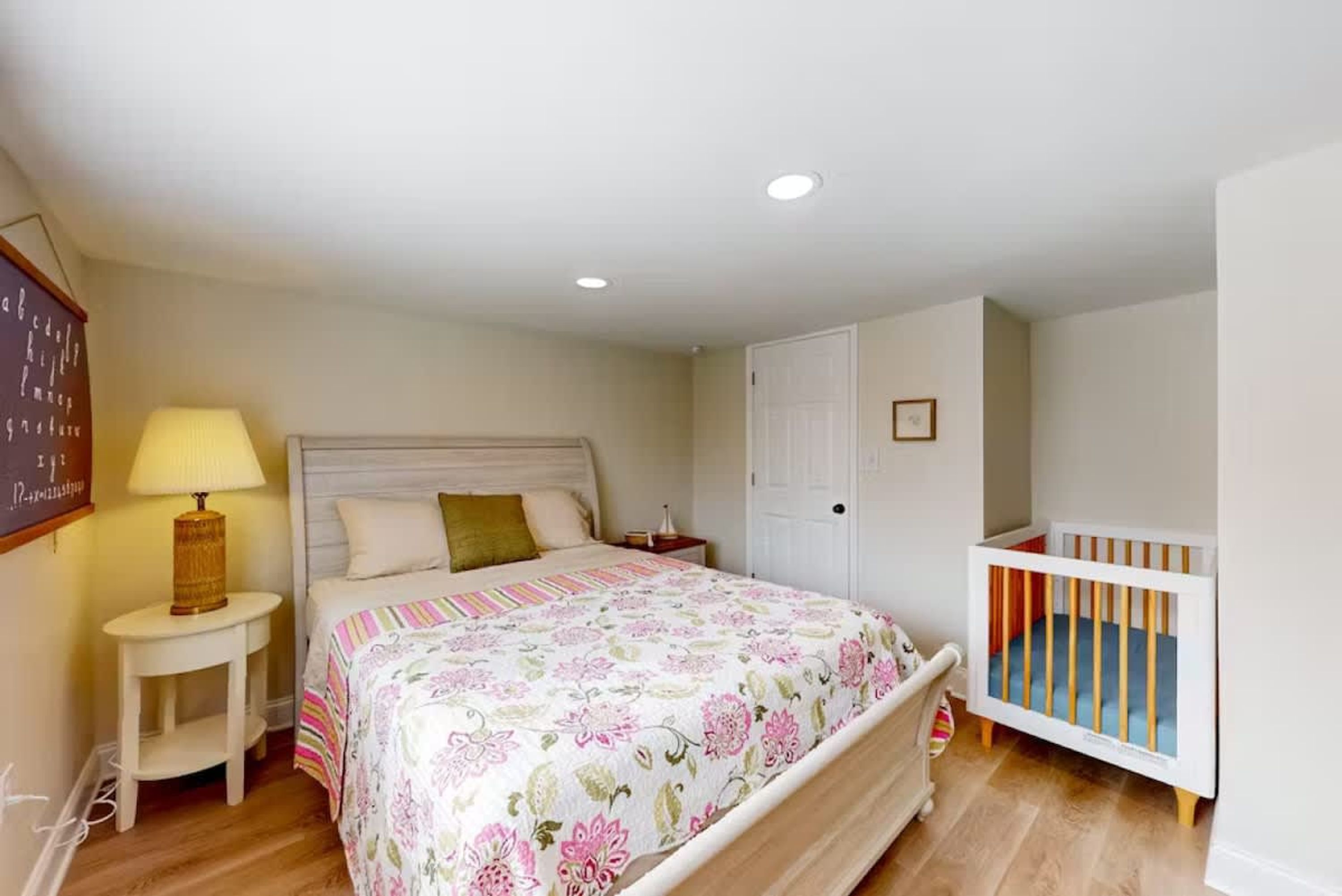 A bedroom with a bed, bedside table, lamp, crib, and a door.