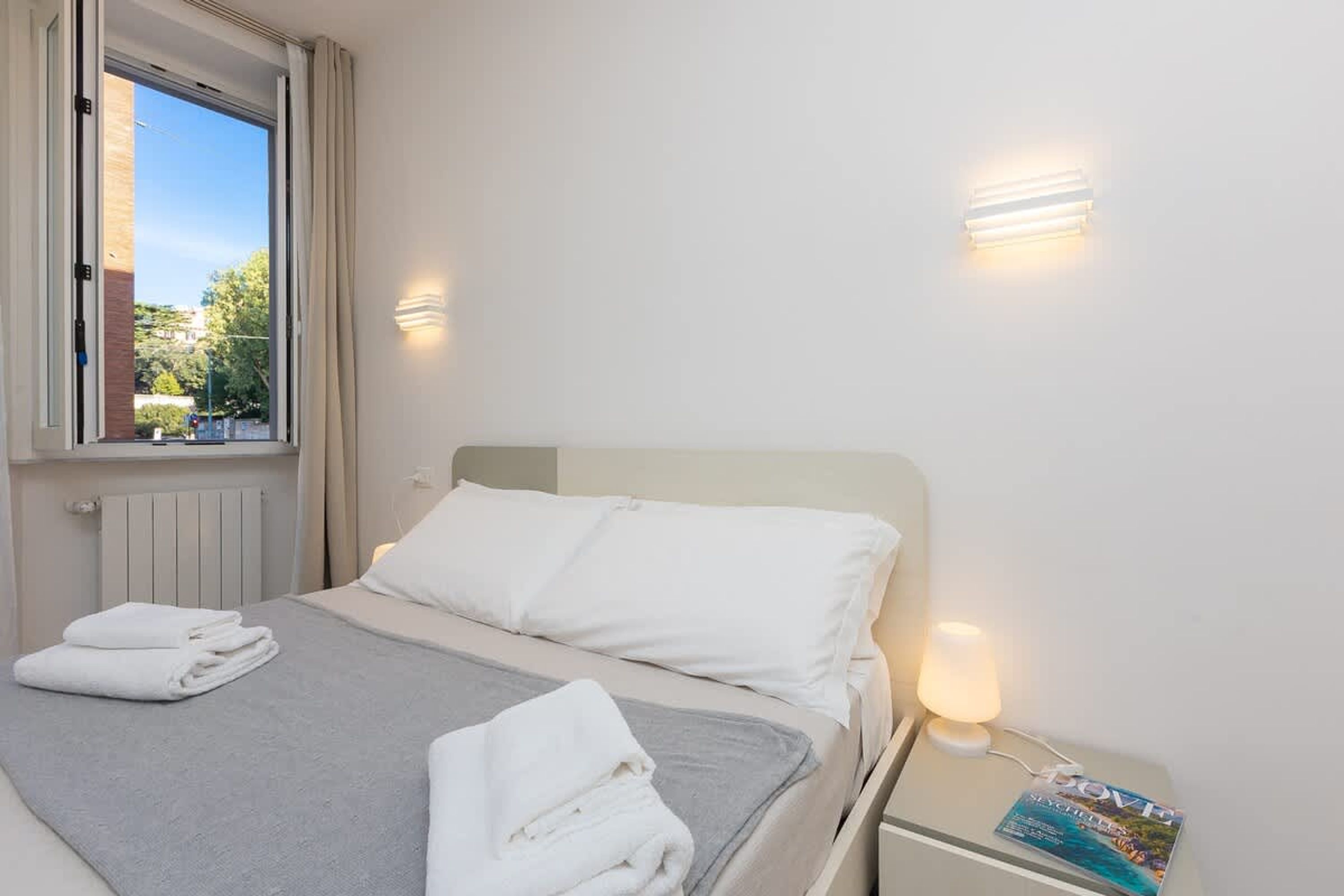 Ein Hotelzimmer mit Bett, Fenster, Heizkörper, Nachttisch mit Lampe und einer Zeitschrift.