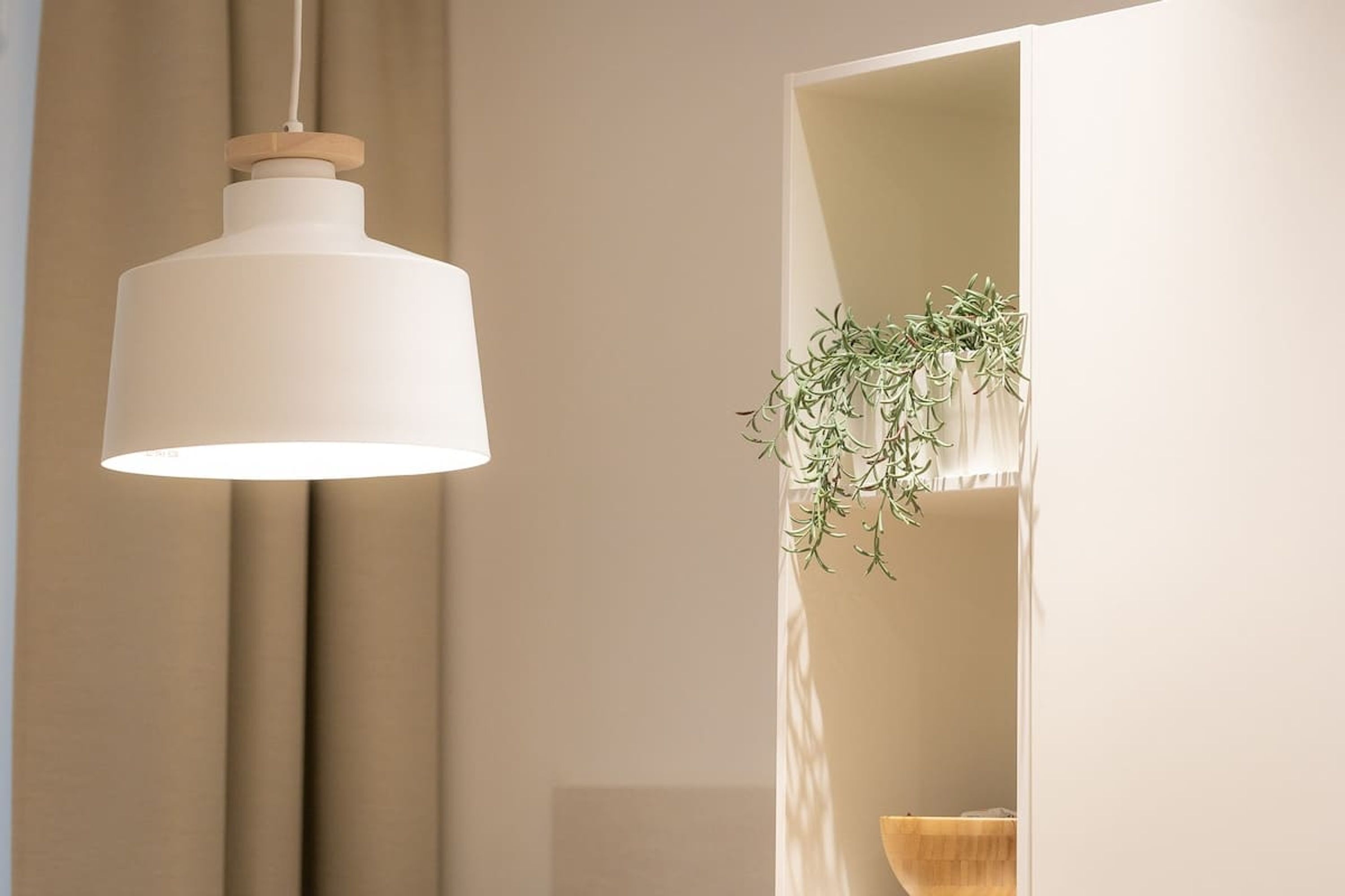 Un luminaire suspendu composé d’un abat-jour blanc et d’une base en bois, installé dans une pièce dont le mur est de couleur beige et où se trouve un bol en bois sur une étagère.
