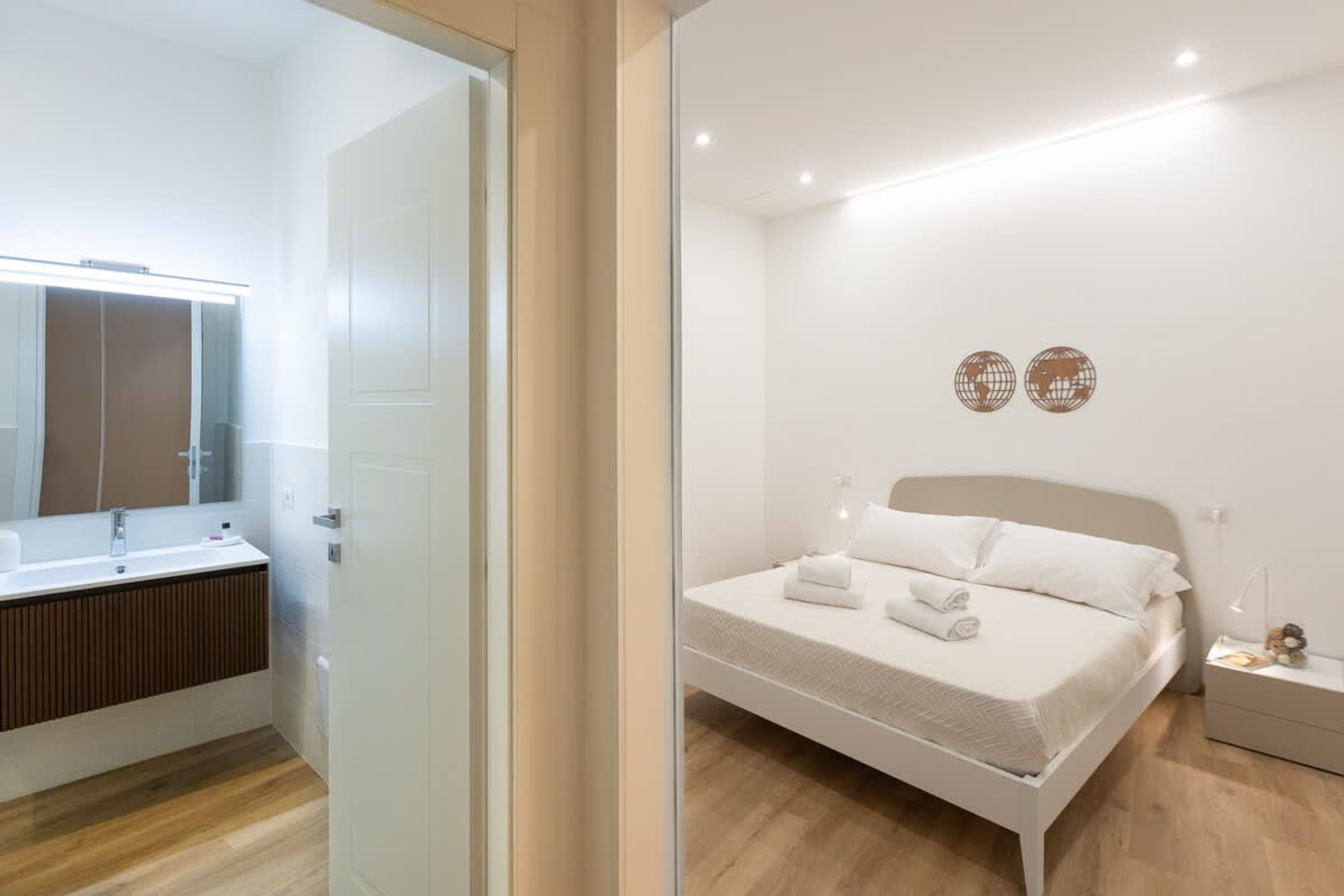A modern bedroom with a white bed, a bedside table, and a nightstand. There is a large mirror above the sink in the adjacent bathroom.