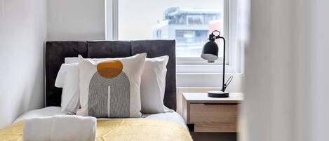 A neatly made bed with a yellow bedspread, a black headboard, and white pillows. A white towel is folded at the foot of the bed. There is a small bedside table with a lamp and a cup. A window with a view of buildings is visible in the background.