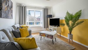 Una sala de estar moderna con un sofá gris, cojines amarillos, una mesa de centro blanca, una planta en maceta y un televisor sobre un mueble blanco.