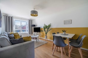 Una sala de estar moderna con un sofá, una mesa de comedor y sillas. Un televisor está montado en la pared, y hay un letrero que dice "THIS IS OUR HAPPY PLACE".