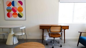 Un espace de bureau moderne avec une table ronde en bois, un bureau blanc, un fauteuil de bureau en osier et un tableau abstrait aux couleurs vives accroché au mur.