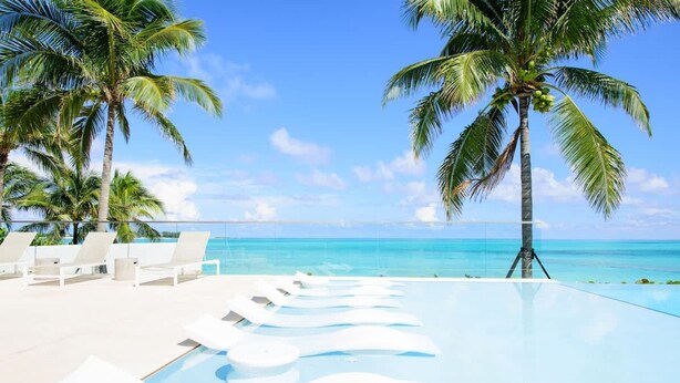 Zona de piscina con sillas reclinables y palmeras, con vista a un océano azul cristalino.