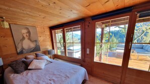 Une chambre de style cabane en bois, avec un grand lit, de nombreux oreillers, un tableau au mur et de grandes fenêtres donnant sur une terrasse et des montagnes.