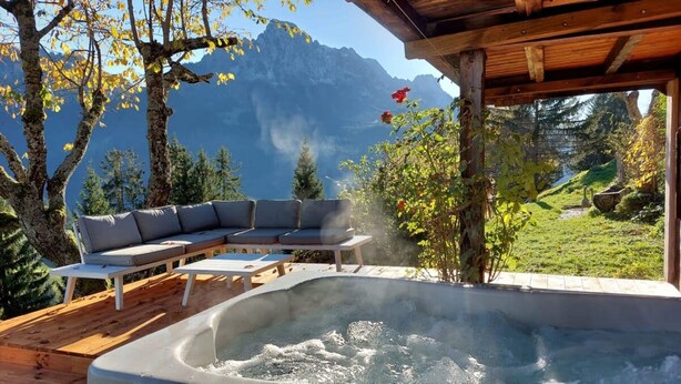 Un bain à remous avec une terrasse en bois et un canapé, entouré d’arbres et de montagnes.