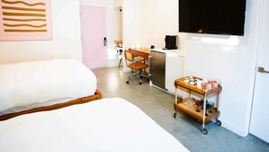Habitació d'hotel amb llit, escriptori, nevera, carretó amb vasos i televisió penjada a la paret.