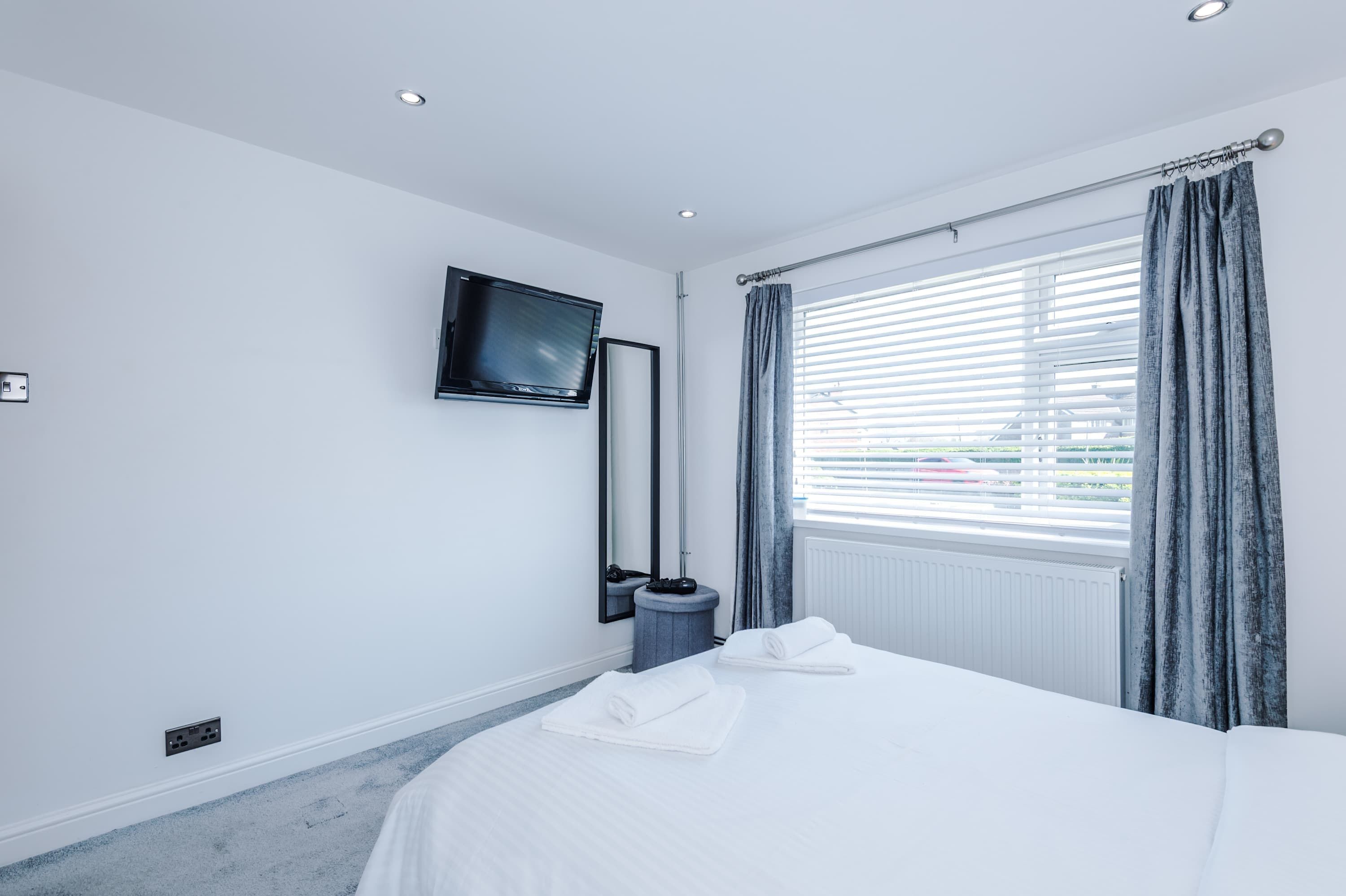 A hotel room with a bed, a flat-screen TV mounted on the wall, a window with blinds, and a radiator.