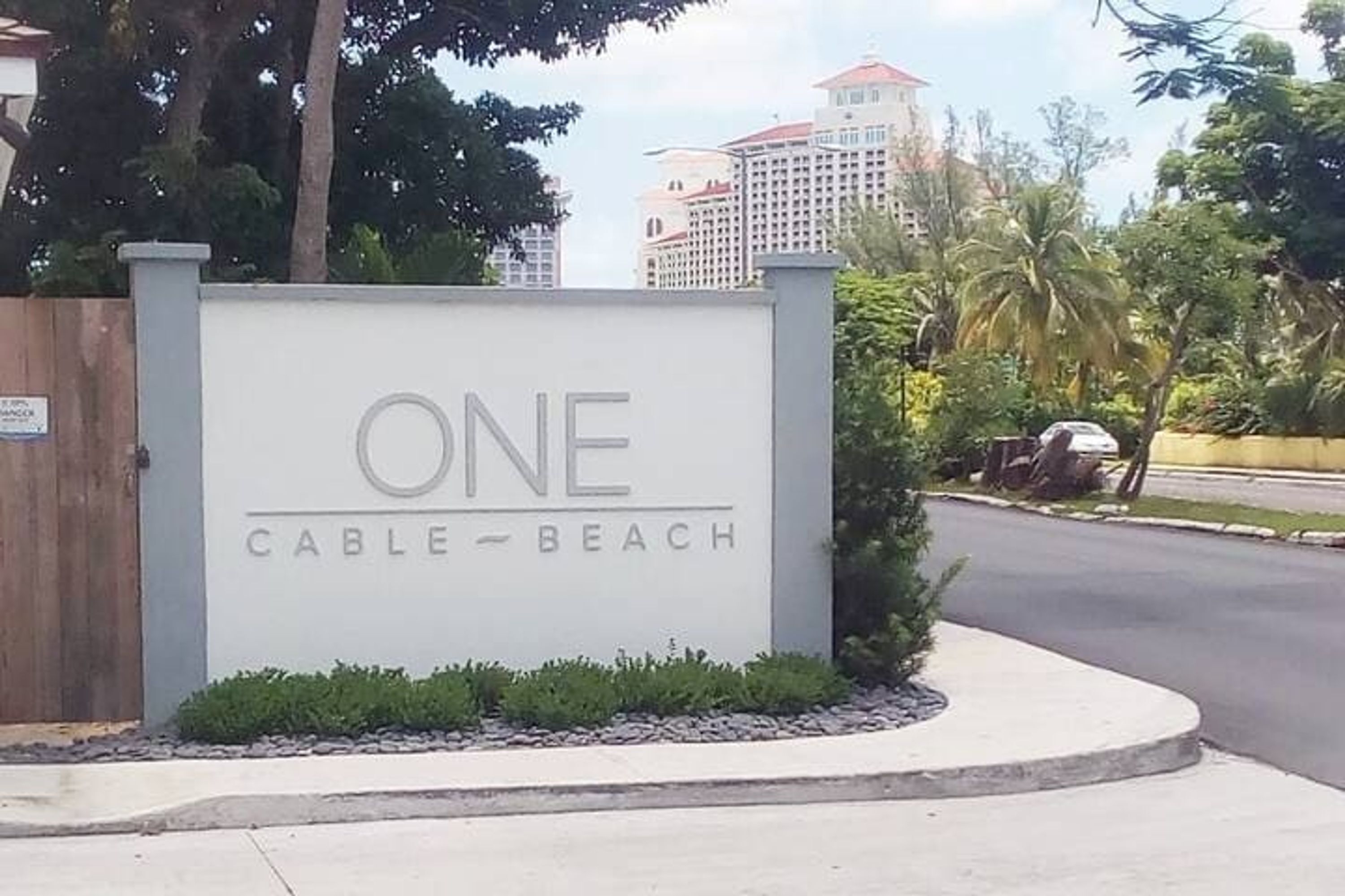 Une enseigne indiquant « ONE Cable-Beach », avec une entrée fermée par un portail, un espace aménagé avec de petites plantes et une route en arrière-plan.