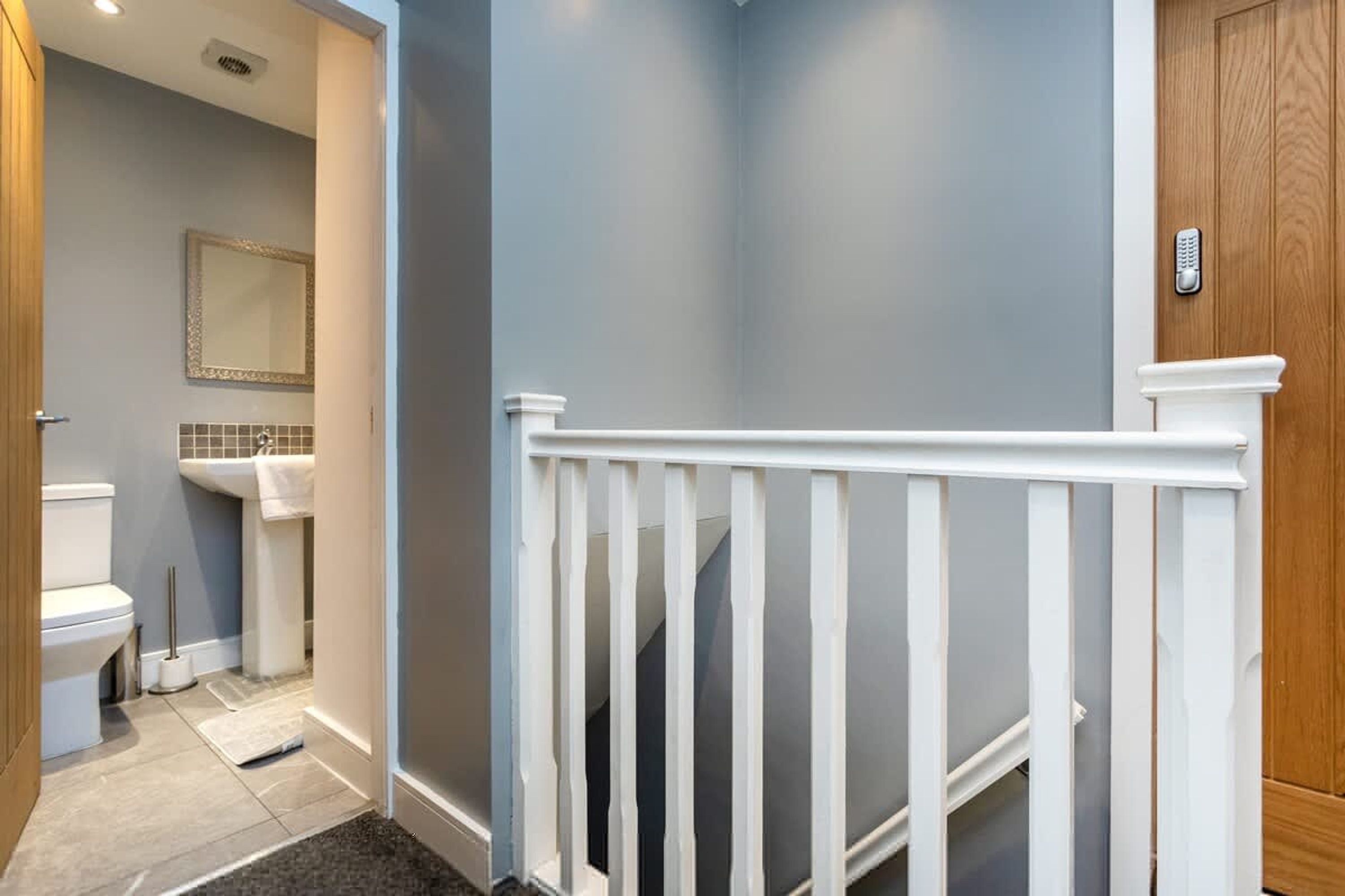 A hallway with a white railing, a wooden door, and a bathroom visible in the background.