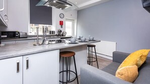 A modern kitchen with a central island, bar stools, and a grey sofa with yellow cushions. - Stylish Affordable Home in Burnley (Burnley)