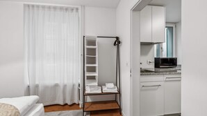 A small, modern room with a bed, a white wardrobe, a kitchen area with a microwave, and a shelf unit with folded towels. - Modern 2 Bdr-flat top Location (Kriens)
