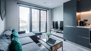 A modern living room with a sofa, a coffee table, and a TV. There is a large window providing a view of buildings outside. - Stylish City Centre Apartment (Leeds)